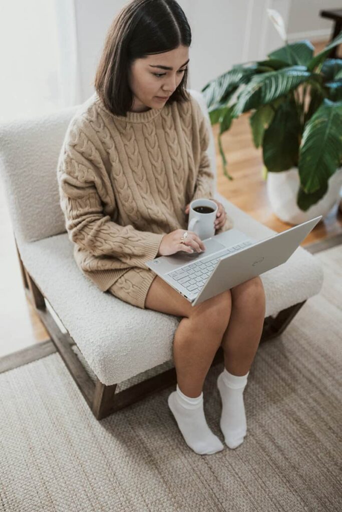 Woman using laptop and drinking coffee in a cozy living room setting.